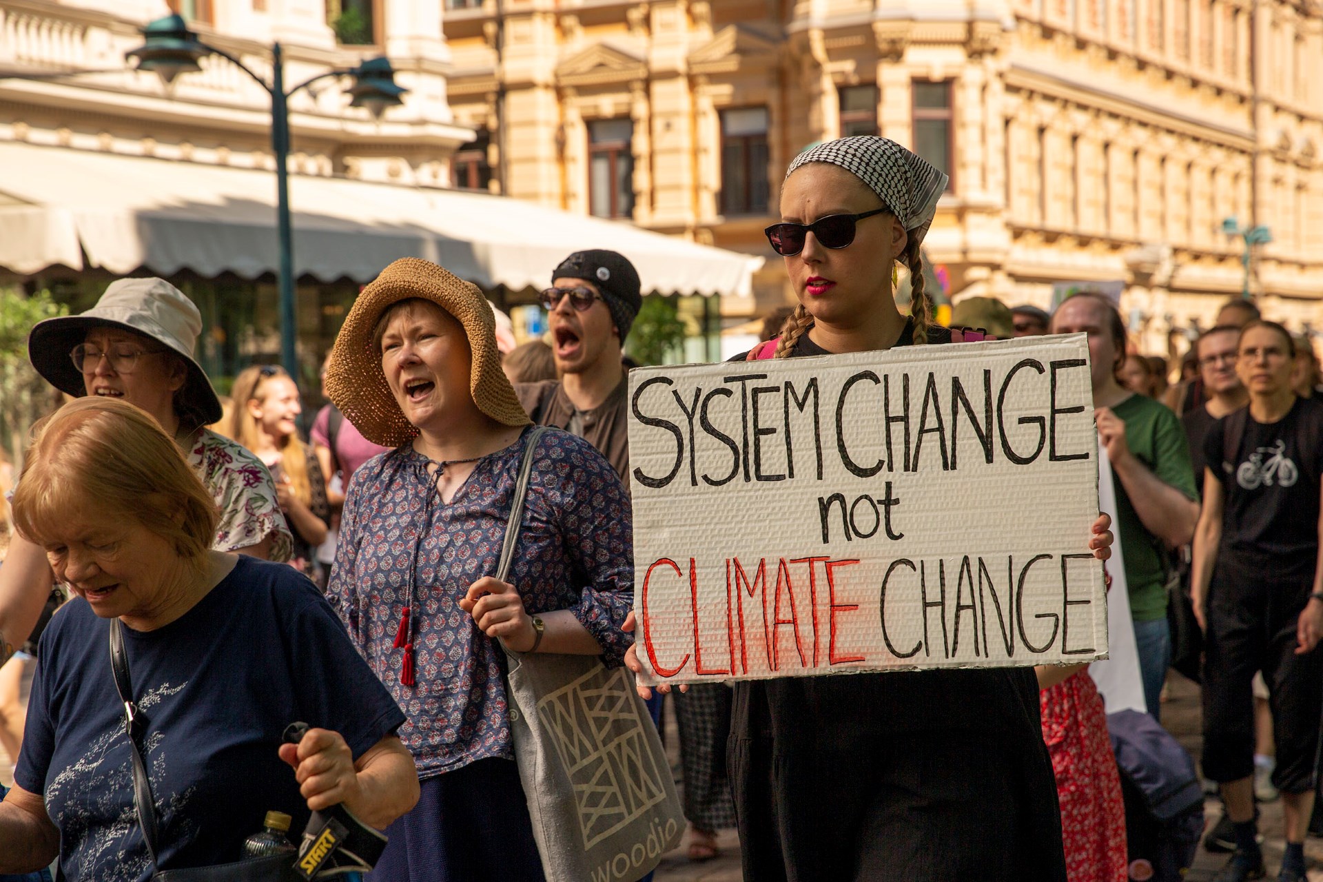 A photo of a group of climate protestors advocating for the preservation of the forest ecosystem. A woman holding a sign saying "System Change Not Climate Change".