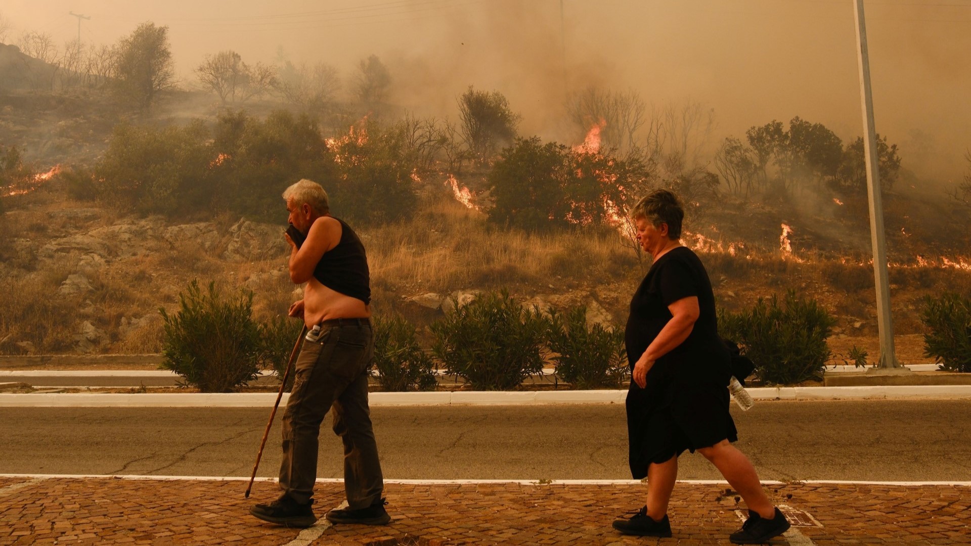 Photo of two people walking and passing by the wildfire in Chasia in the outskirts of Athens.