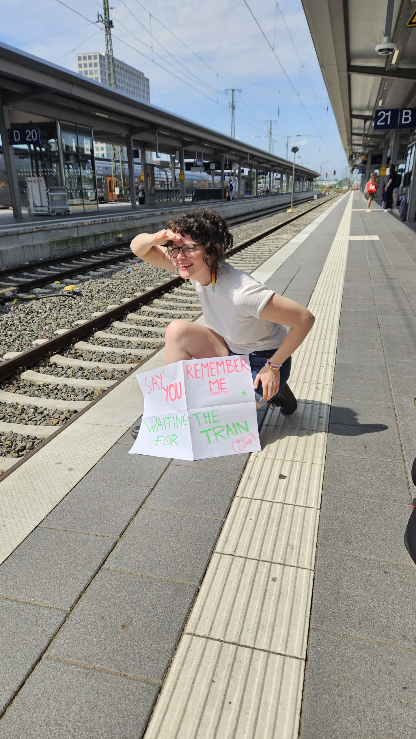 Clara Francken at a train station. No train in sight. She's holding a sign that reads, "Say You Remember Me / Waiting for the Train."