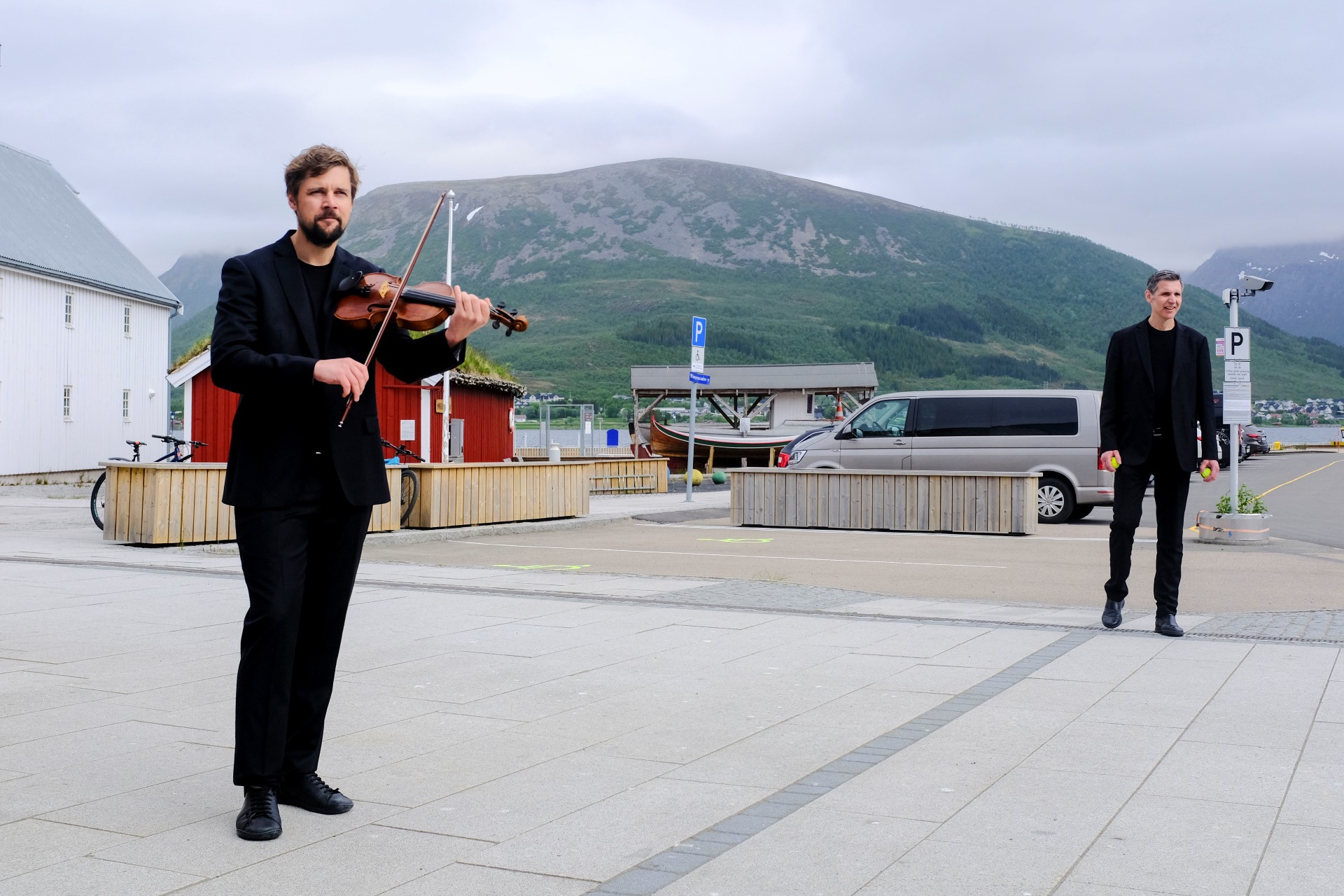 Two men in suits on an open street in Sortland, Norway. One is holding a violin while the other is ready to start joggling. 