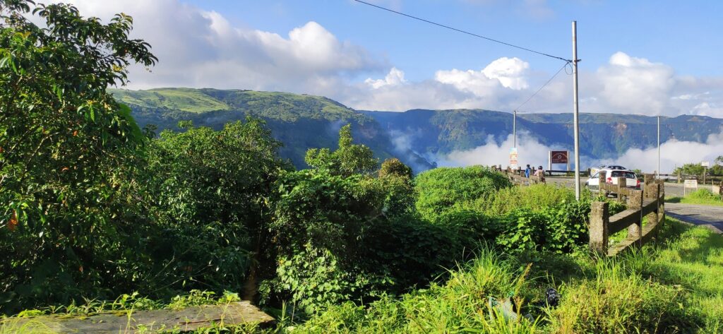 A photo of a street in Meghalaya. A single car and a couple of pedestrians next to a wide landscape of mountains, forests, and nature