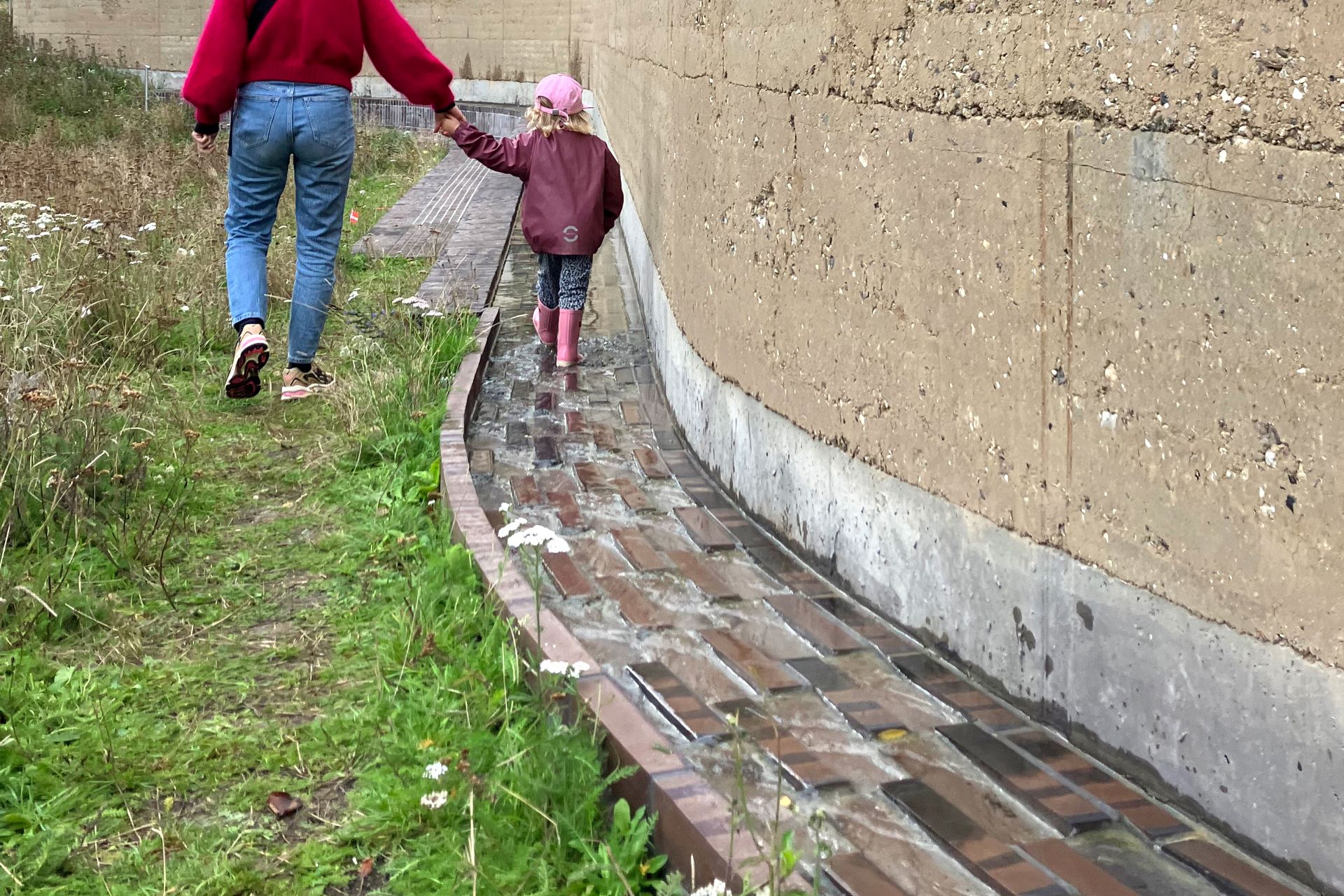 A photo of a child holding hands with an adult while walking along the ‘climate wall’, and playing in the puddles.